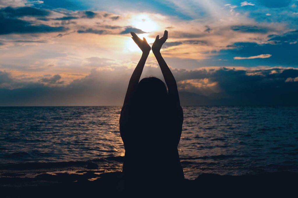 Shadow of woman with arms up toward sky standing in front of water as sun sets